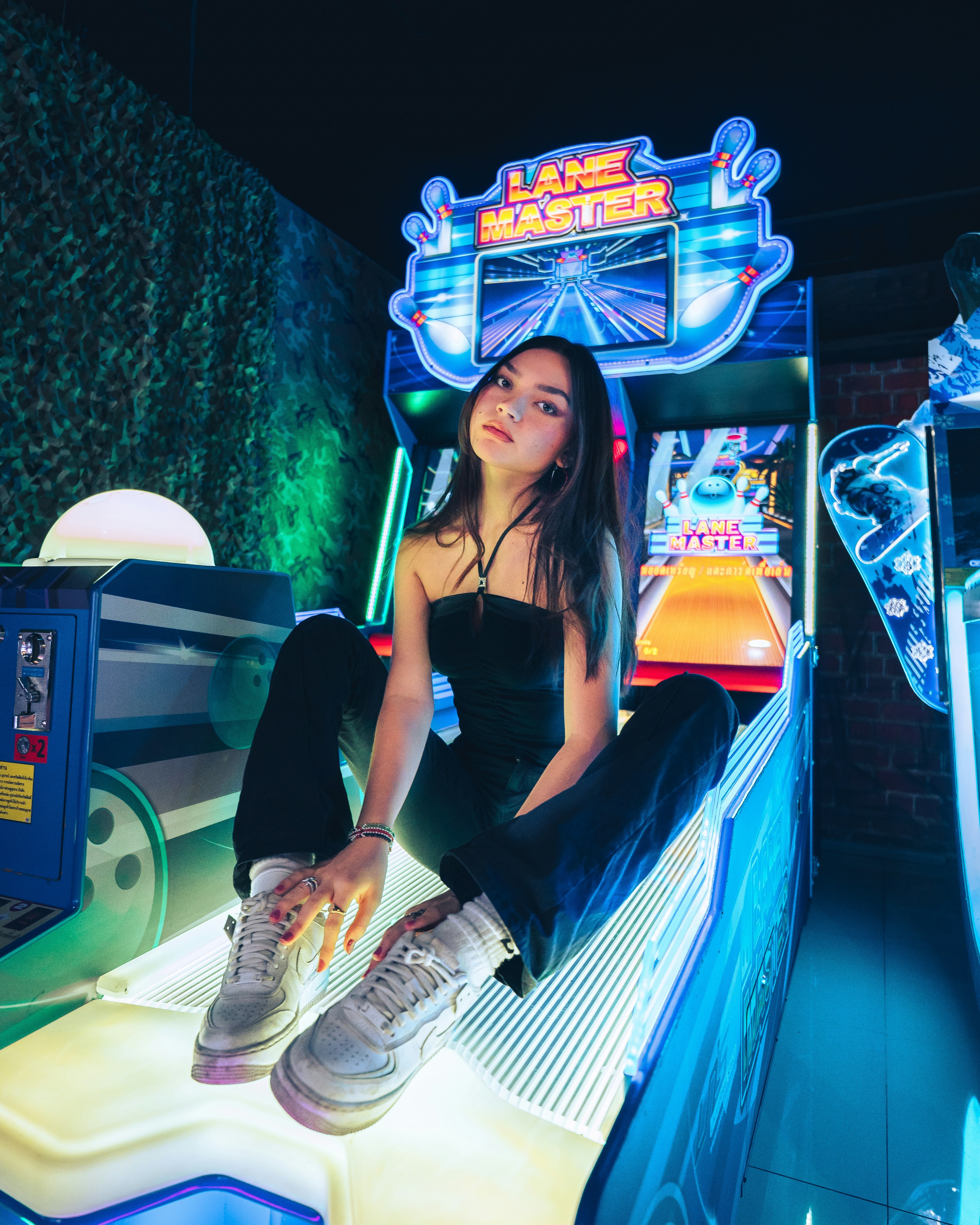 A person sitting on an arcade game machine platform with neon lights in the background. The arcade game is called 'Lane Master' and features a colorful design with bowling-themed imagery.