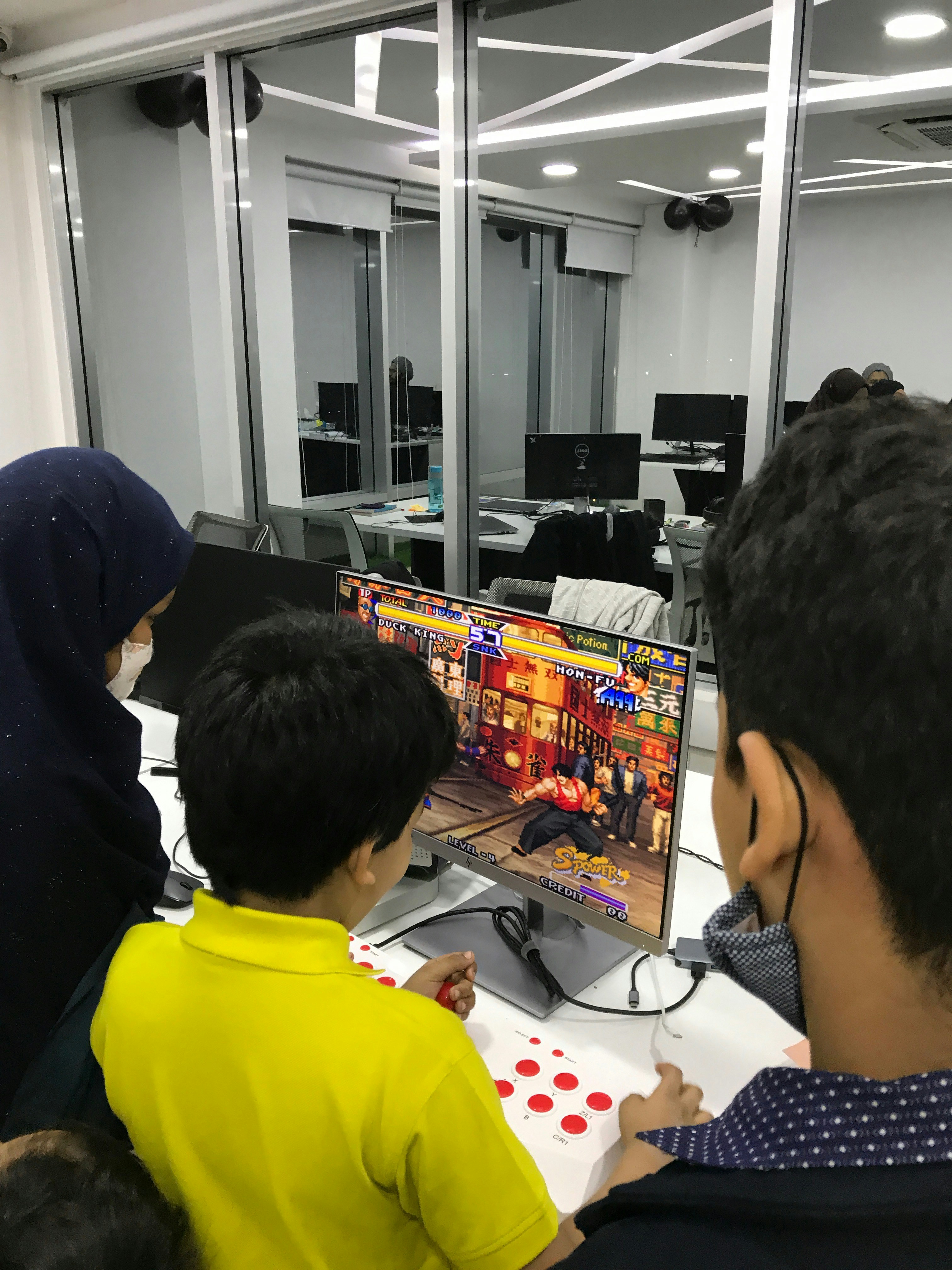 A group of children is gathered around a desktop computer, engaged in playing a colorful, action-packed arcade game displayed on the monitor. The setting appears to be a modern, well-lit office or gaming space with large glass windows, showing multiple computer workstations in the background.