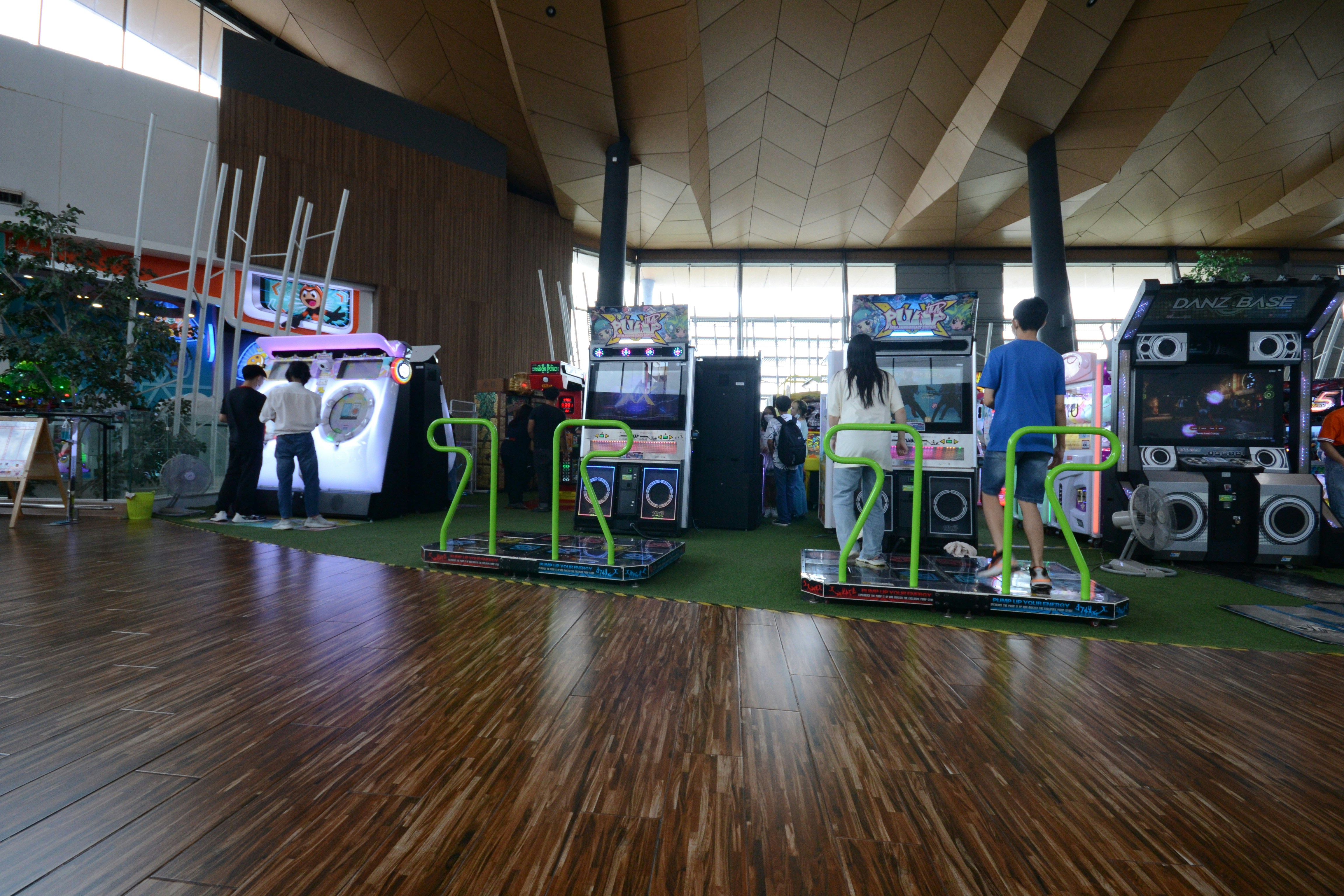 A vibrant arcade setting with several gaming machines lined up, including dance and rhythm game setups. People are engaged with the games, absorbed in their activities. The flooring is wooden, and the ceiling has a geometric design. Natural light filters through large windows, creating a lively atmosphere.