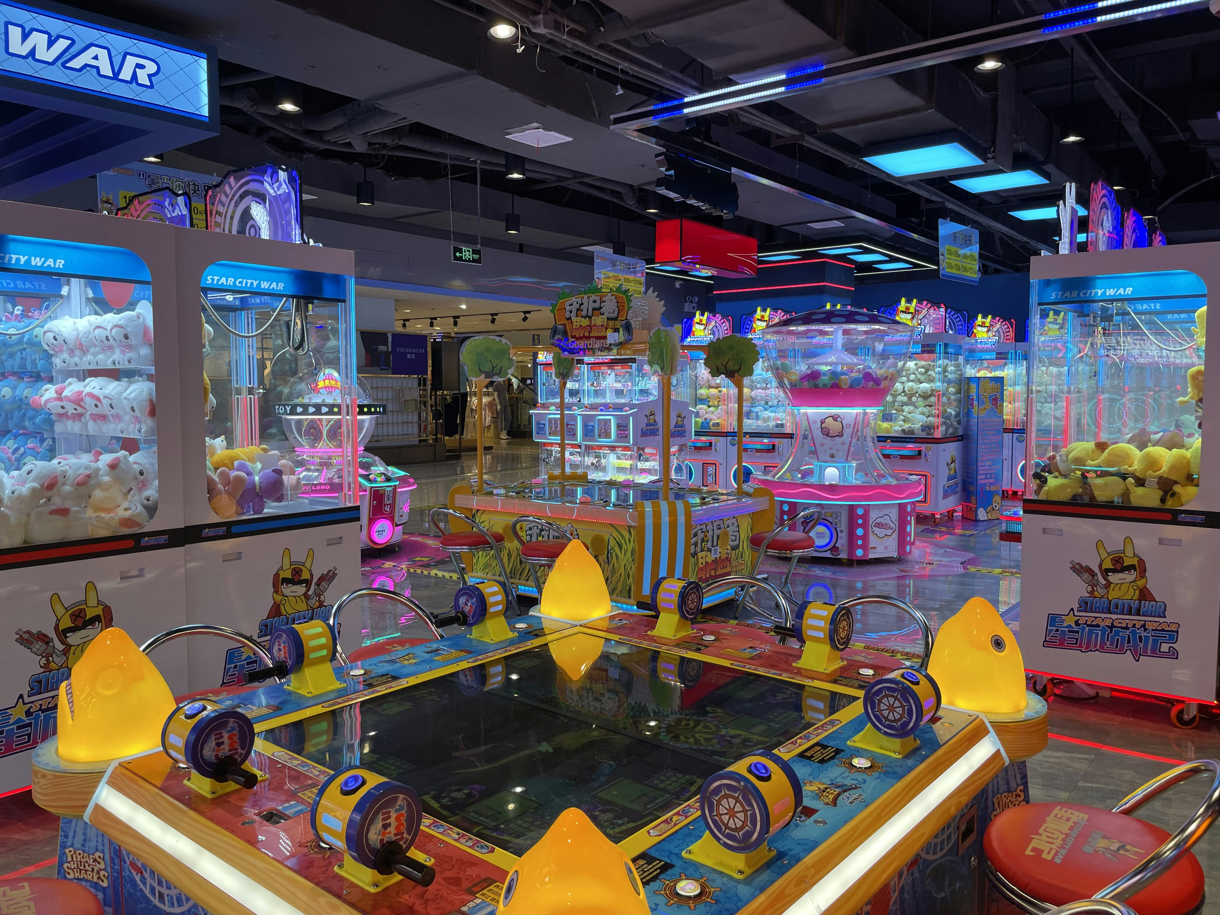 Brightly lit arcade filled with various game machines, including claw machines with stuffed toys, a ticket redemption game, and colorful decorative lighting. The environment is lively and playful, with numerous arcade game consoles lining the floor.